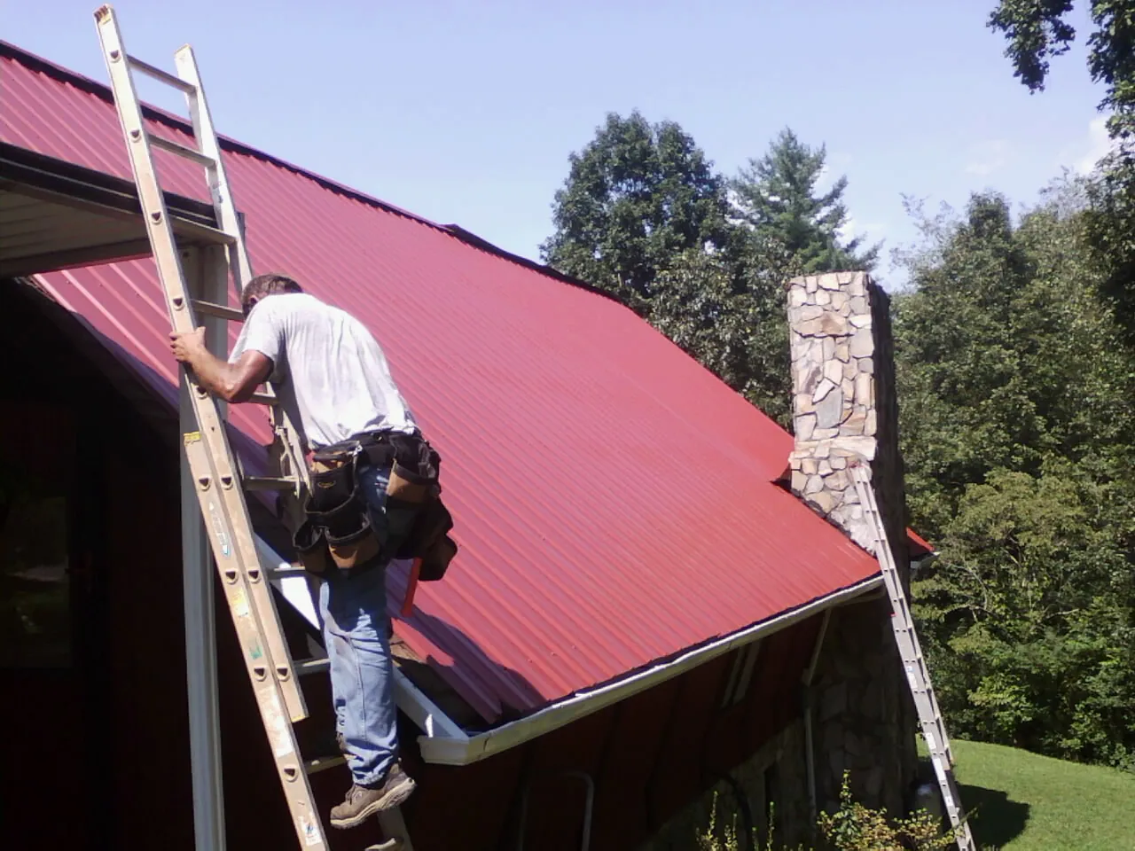 Roofer on a ladder beside a red metal roof and stone chimney — Soffit & Fascia Repair by Roofing Brothers in Maybell